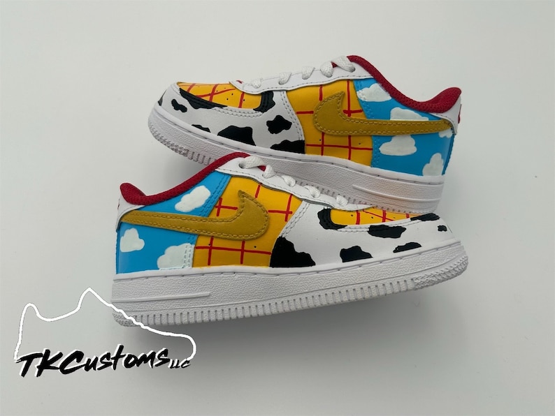 Op de afbeelding: Witte, gele, rode en blauwe custom-gekleurde sneakers met een koe-print ontwerp. De sneakers hebben een rood swoosh-logo en een blauw ontwerp met witte wolken aan de zijkant.