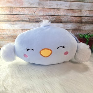 May include: A light blue plush bird pillow with a yellow beak, pink cheeks, and embroidered eyes. The pillow has white pom-poms on each side.
