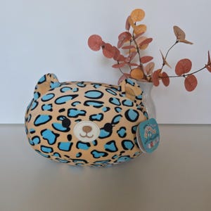 May include: A plush, round leopard-print pillow with a light brown base and blue spots. The pillow has a bear-like face with a tan nose and a tag attached. A vase with orange and brown leaves is in the background.