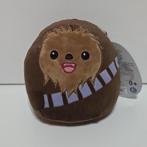 May include: A brown plush toy shaped like Chewbacca, featuring a smiling face with black eyes and a pink mouth. It has a light brown fur design around the face and a light blue bandolier. The toy is on a white surface.