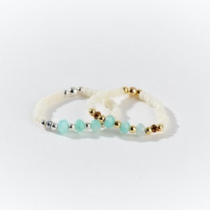 Peut inclure: Deux bagues en perles avec des perles blanches et des accents de pierres précieuses bleu turquoise. La bague à gauche a des perles argentées et la bague à droite a des perles dorées.