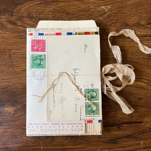 May include: A stack of vintage envelopes tied together with twine and a brown ribbon. The envelopes have stamps and handwritten addresses. The top envelope has the text "AVION AIR MAIL" printed on it. The envelopes are tied with a brown ribbon and twine.