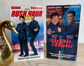 May include: Two VHS tapes of the movie Rush Hour 2, featuring Jackie Chan and Chris Tucker. The tapes are in a vertical orientation, with the left tape showing Chan and Tucker in a fighting pose, and the right tape showing close-up shots of the actors. The text on the tapes reads "Rush Hour 2" and "Hilarious! Better than the first!" on the left tape, and "The year's funniest action-comedy!" on the right tape.