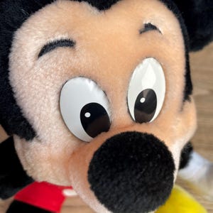 May include: Close-up of a plush Mickey Mouse toy. The toy has a black nose, black ears, and large white eyes with black pupils. The face is a light tan colour, and the toy has a red and black outfit with yellow gloves.