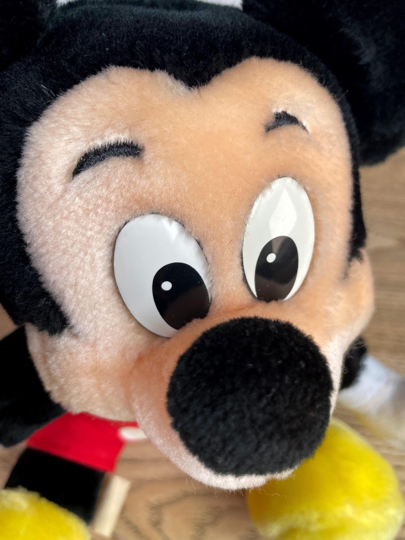 May include: Close-up of a plush Mickey Mouse toy. The toy has a black nose, black ears, and large white eyes with black pupils. The face is a light tan colour, and the toy has a red and black outfit with yellow gloves.
