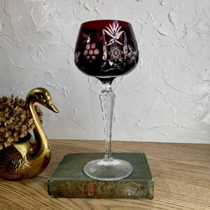 May include: A deep red cut-to-clear crystal wine glass featuring intricate starburst and grape cluster engravings on the bowl. The stem is faceted and clear, leading down to a circular base. The glass displays a rich ruby color with clear glass.