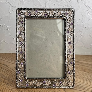 May include: A rectangular silver-toned metal picture frame featuring an intricate openwork border decorated with light pink enamel flowers and small, sparkling purple crystals embedded within the floral and vine-like metal design.
