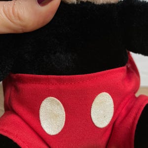 May include: A plush Mickey Mouse toy, with a black body, tan face, and red shorts with white circular buttons. The toy's design is a classic representation of the iconic cartoon character. The plush material gives it a soft, huggable appearance.