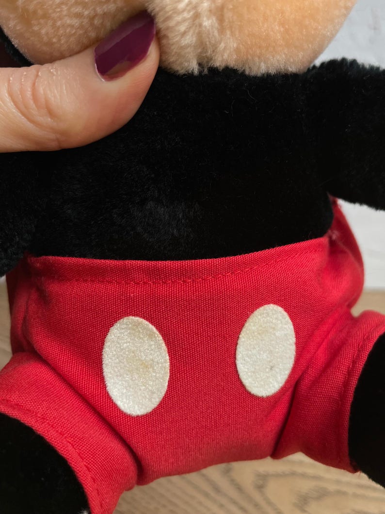 May include: A plush Mickey Mouse toy, with a black body, tan face, and red shorts with white circular buttons. The toy's design is a classic representation of the iconic cartoon character. The plush material gives it a soft, huggable appearance.