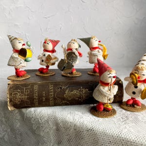 May include: A collection of vintage Christmas ornaments depicting a musical band. Each figure is dressed in white, red, and gold, with instruments including a drum, violin, and saxophone. The ornaments are displayed on a vintage book.