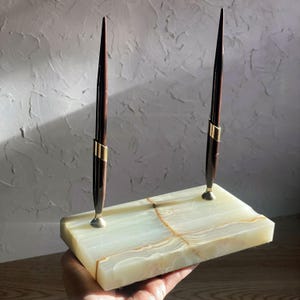 May include: A pen holder with two dark brown pens with gold accents. The pens are set in a rectangular, light green and brown marble base. The pen holder is a stylish desk accessory.