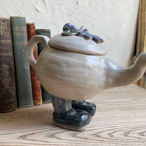 May include: A white ceramic teapot with a blue and green floral design on the lid. The teapot has legs and black shoes.