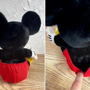 May include: A plush Mickey Mouse doll, seen from the rear, with large black ears, a black body, and red shorts. The doll has white gloves and yellow accents. The doll is in a sitting position.