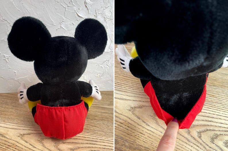 May include: A plush Mickey Mouse doll, seen from the rear, with large black ears, a black body, and red shorts. The doll has white gloves and yellow accents. The doll is in a sitting position.