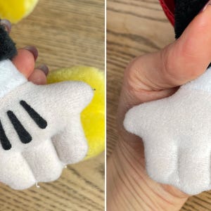 May include: Close-up of a plush toy hand, white with three black lines on the palm. The hand is part of a larger character, with a black sleeve and a red accent visible. The toy is soft and likely made of felt or similar material.