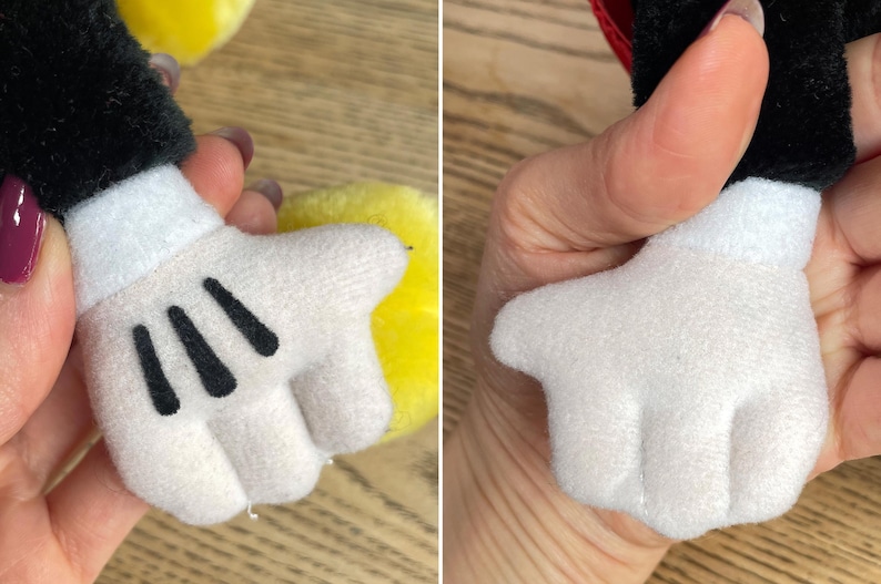 May include: Close-up of a plush toy hand, white with three black lines on the palm. The hand is part of a larger character, with a black sleeve and a red accent visible. The toy is soft and likely made of felt or similar material.