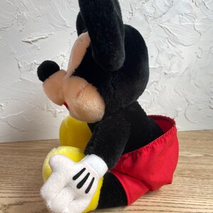 May include: A plush Mickey Mouse toy, with black ears, head, and a tan face. The toy wears red shorts, white gloves with black stripes, and yellow shoes. The toy is in a sitting position.