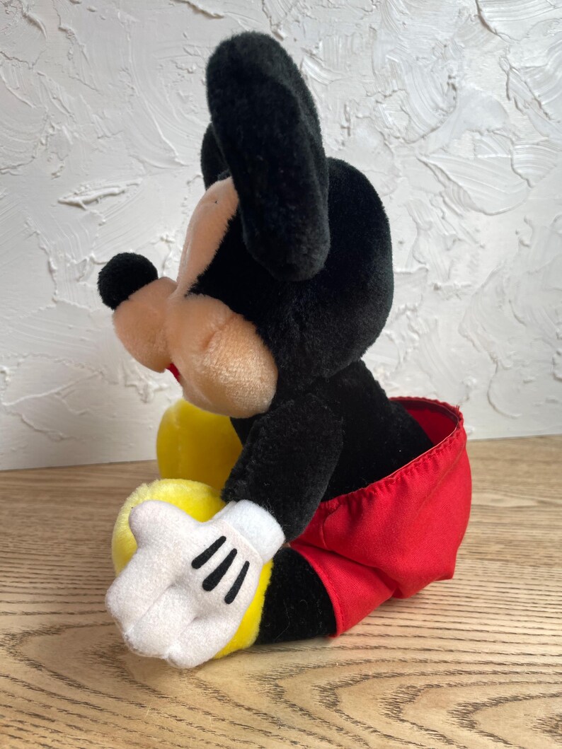 May include: A plush Mickey Mouse toy, with black ears, head, and a tan face. The toy wears red shorts, white gloves with black stripes, and yellow shoes. The toy is in a sitting position.