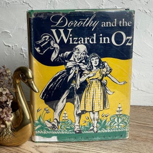 May include: A hardcover book titled Dorothy and the Wizard in Oz by L. Frank Baum. The cover features a black, yellow, and green illustration of a man in a top hat and a girl in a checkered dress standing in a field of stylized green plants.