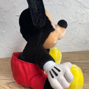 May include: A plush Mickey Mouse toy, with black fur, a tan face, and a black nose. The toy wears red shorts, white gloves with black details, and yellow shoes. The classic character is in a seated position.