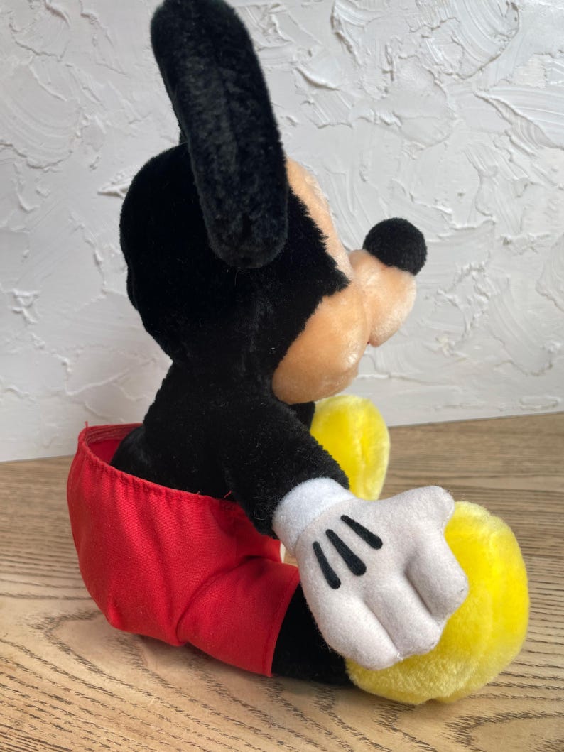 May include: A plush Mickey Mouse toy, with black fur, a tan face, and a black nose. The toy wears red shorts, white gloves with black details, and yellow shoes. The classic character is in a seated position.