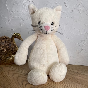 May include: A plush cat toy with soft cream-colored fur, featuring a small pink nose, embroidered black eyes, and long black whiskers. The stuffed animal has a rounded body, long arms, and short legs, sitting in an upright position with a gentle expression.