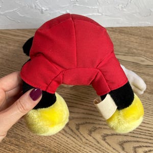 May include: A plush toy with a red body, black legs, and yellow feet. The toy has white gloves and black ears. The toy is a classic cartoon character.
