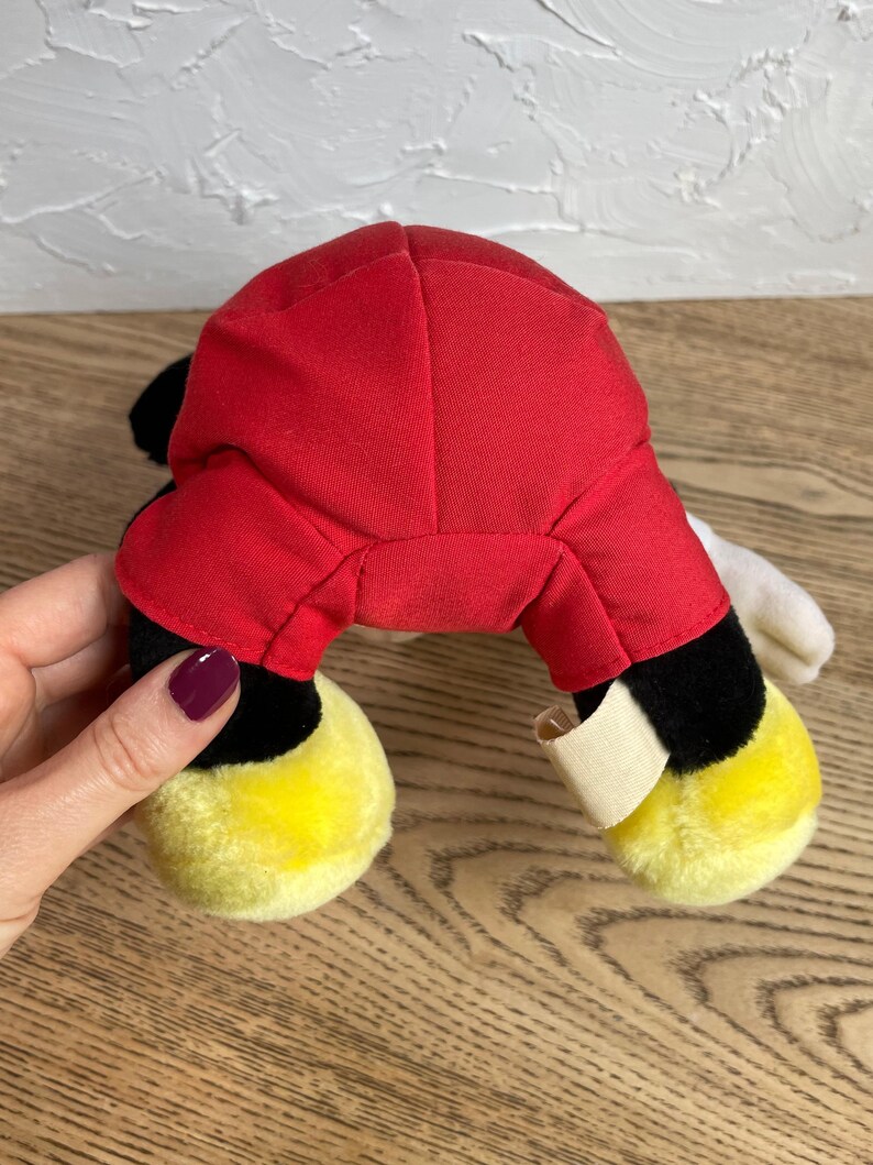 May include: A plush toy with a red body, black legs, and yellow feet. The toy has white gloves and black ears. The toy is a classic cartoon character.