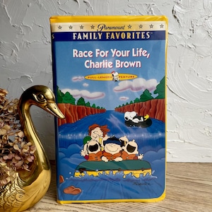 May include: A yellow VHS tape with a cartoon image of Charlie Brown and Snoopy on a raft going down a river. The title of the tape is "Race For Your Life, Charlie Brown" and it is a Paramount Family Favorites full-length feature.