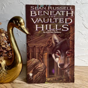 May include: A hardcover book titled "Beneath the Vaulted Hills" by Sean Russell, featuring a wolf illustration. The book is part of "The River Into Darkness" series. A gold swan figurine is on the left.