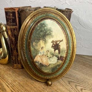 May include: Oval-shaped vintage wall art with a gold and teal frame. The artwork depicts a scene with three figures and a musician playing a lute. The frame has a decorative gold trim. The artwork is displayed on a wooden surface.