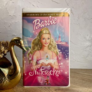 May include: A VHS tape of the movie Barbie in the Nutcracker featuring a blonde character wearing a gold crown and a pink dress. The cover art displays the title Barbie in the Nutcracker with a castle in the background and a small nutcracker figure.