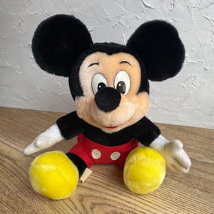 May include: A plush Mickey Mouse doll with black ears, a black nose, and a smiling face. Mickey wears a red pair of shorts with white buttons, black gloves, and yellow shoes. The stuffed animal is sitting on a wooden surface.