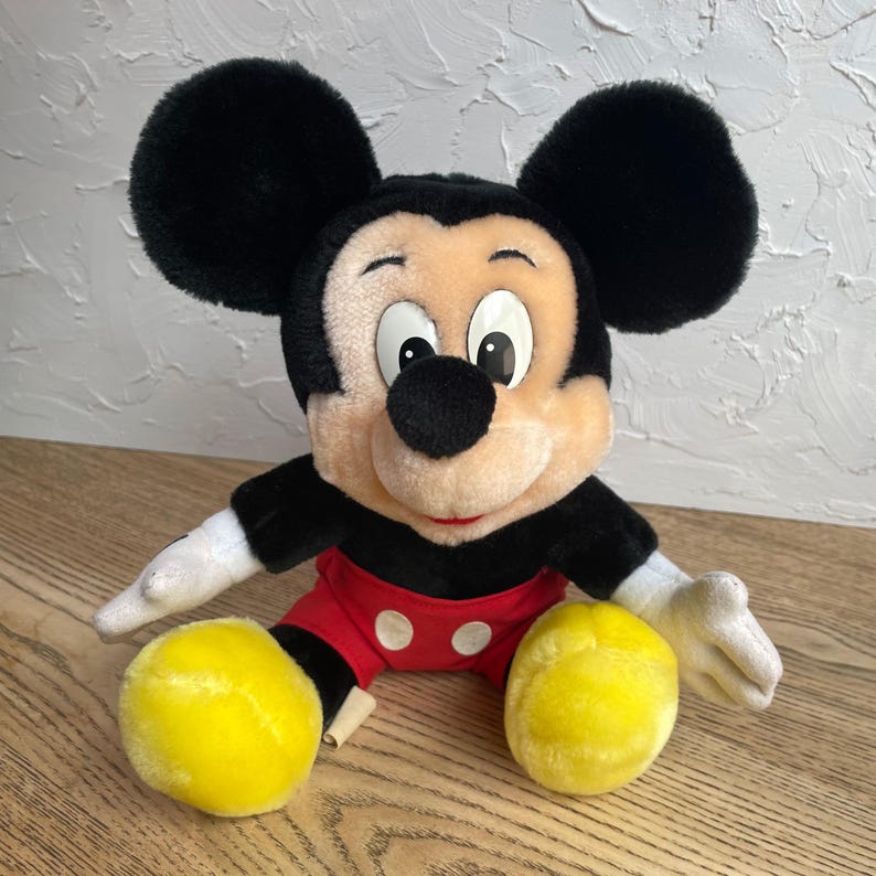 May include: A plush Mickey Mouse doll with black ears, a black nose, and a smiling face. Mickey wears a red pair of shorts with white buttons, black gloves, and yellow shoes. The stuffed animal is sitting on a wooden surface.