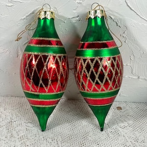Puede incluir: Dos adornos navideños con forma de lágrima. Los adornos son verdes y rojos con detalles dorados brillantes. La parte superior e inferior son verdes, con una banda roja en el medio. Un patrón de diamantes dorados brillantes cubre la sección roja.