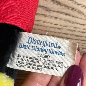 May include: A close-up of a tag from a Disneyland Walt Disney World plush toy. The tag is rectangular with the words "Disneyland Walt Disney World" and "&copy;Disney" printed on it. The tag also includes the text "ALL NEW MATERIALS POLYESTER FIBERS".