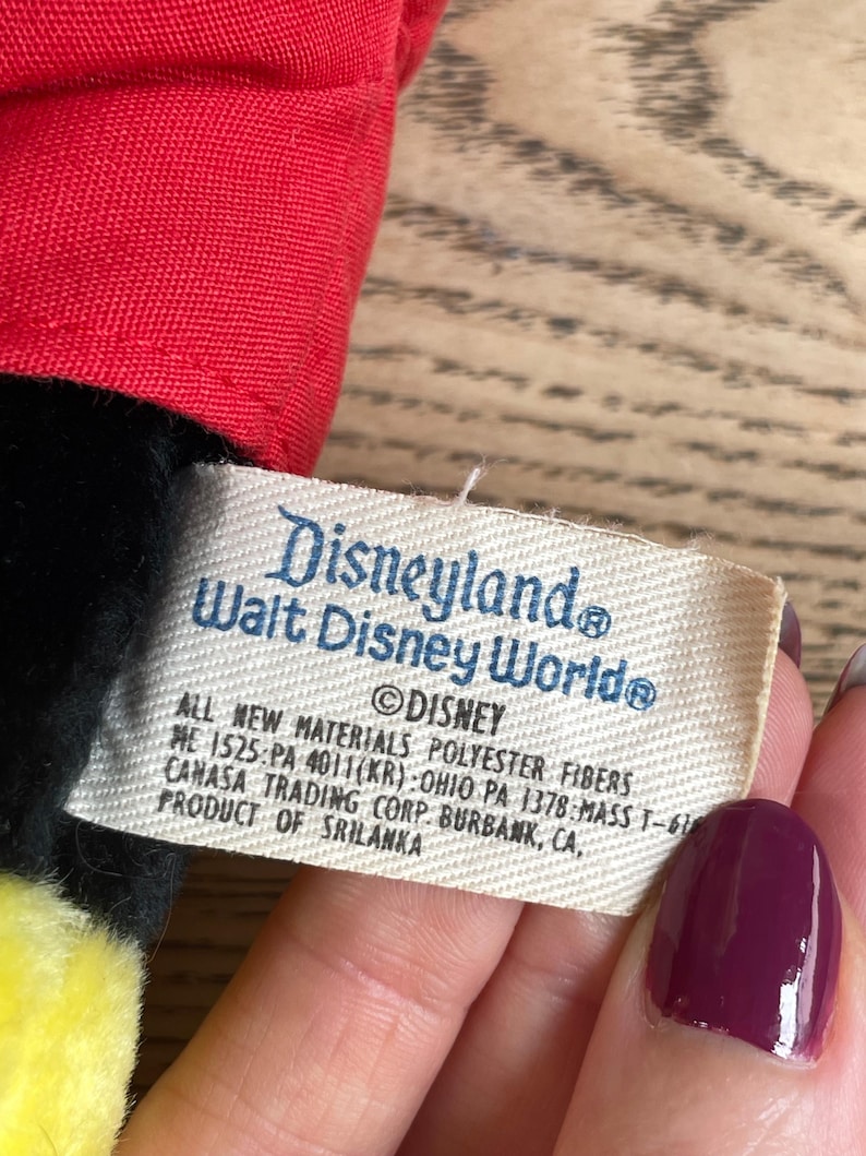 May include: A close-up of a tag from a Disneyland Walt Disney World plush toy. The tag is rectangular with the words "Disneyland Walt Disney World" and "&copy;Disney" printed on it. The tag also includes the text "ALL NEW MATERIALS POLYESTER FIBERS".
