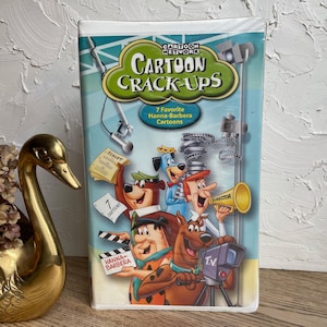 May include: A VHS tape featuring a cartoon illustration of characters from Hanna-Barbera cartoons. The title is "Cartoon Crack-Ups" with the tagline "7 Favorite Hanna-Barbera Cartoons".