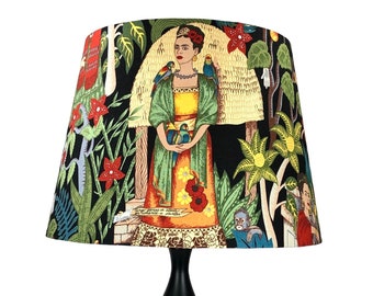 May include: Black table lamp with a fabric lampshade featuring a colorful illustration of Frida Kahlo surrounded by tropical plants and birds. The lampshade has a black background and the illustration is in shades of green, yellow, orange, red, and blue.
