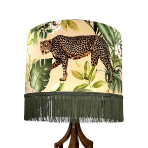 Puede incluir: Una pantalla de lámpara beige con flecos verdes presenta un estampado de leopardo y hojas tropicales. La pantalla está sobre una base de lámpara de madera.