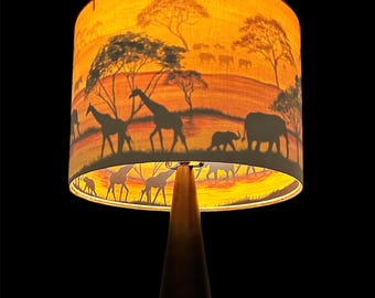 Könnte beinhalten: Dekorative Lampe mit konischem Holzfuß und Lampenschirm mit einer Savannenszene. Der Lampenschirm zeigt Silhouetten von Giraffen, Elefanten und Bäumen vor einem warmen orange-gelben Hintergrund, wodurch ein Sonnenuntergangseffekt entsteht.