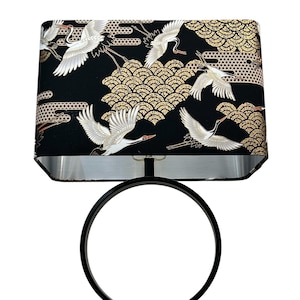Puede incluir: Lámpara de mesa negra con una pantalla rectangular con un diseño de grullas japonesas en blanco y dorado sobre un fondo negro. La lámpara tiene una base circular de metal negro y una base cuadrada.