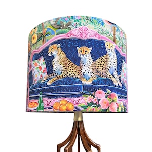 May include: A drum-shaped lampshade featuring a vibrant print of three cheetahs resting on a dark blue tufted sofa. The design includes pink ornate trim, floral accents, green foliage, a green bottle, and orange fruit against a colorful background.