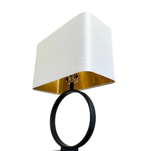 May include: A table lamp featuring a rectangular white fabric shade with a metallic gold interior lining. The lamp base consists of a matte black circular frame supported by a solid rectangular black base, with a central vertical stem.