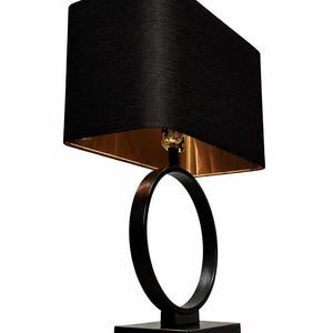 Puede incluir: Una lámpara de mesa negra moderna con una pantalla rectangular y un interior dorado. La lámpara presenta un elemento de diseño circular y una base cuadrada. La lámpara está diseñada para uso en interiores.