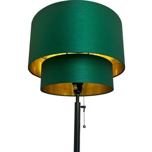 May include: A floor lamp featuring a double-tiered drum lampshade with a textured dark green exterior and a metallic gold interior lining. The lamp has a slender black metal pole and includes a silver pull chain switch for operation.