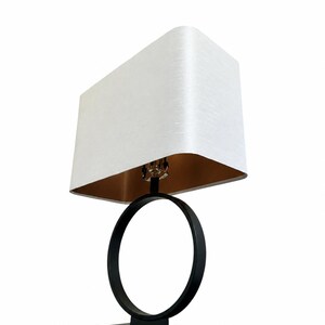 Può includere: Una lampada da tavolo con un paralume rettangolare in tessuto bianco e un rivestimento interno color oro metallizzato. La base della lampada è composta da una cornice circolare nera su un supporto rettangolare nero, con un'asta centrale nera.