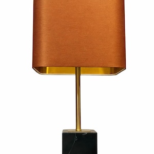 Puede incluir: Una lámpara de mesa moderna con una pantalla rectangular de color óxido. La lámpara presenta un vástago y una base dorados, y una base cúbica de mármol negro. La lámpara está diseñada para uso en interiores.