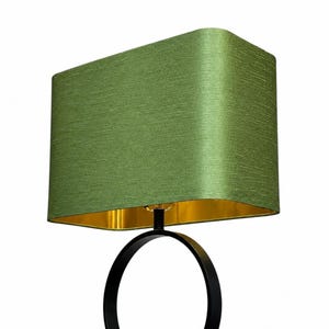 May include: A table lamp featuring a rectangular green fabric lampshade with a textured finish and a metallic gold interior lining. The base consists of a thin black metal frame shaped into a circular ring that supports the lampshade from below.