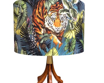 Könnte beinhalten: Eine Tischlampe mit einem zylindrischen Lampenschirm, der mit einem Tiger und tropischem Blattwerk in Grün und Schwarz auf dunkelgrauem Grund bedruckt ist, auf einem geschnitzten Holzfuß mit einem messingfarbenen Metallhals.